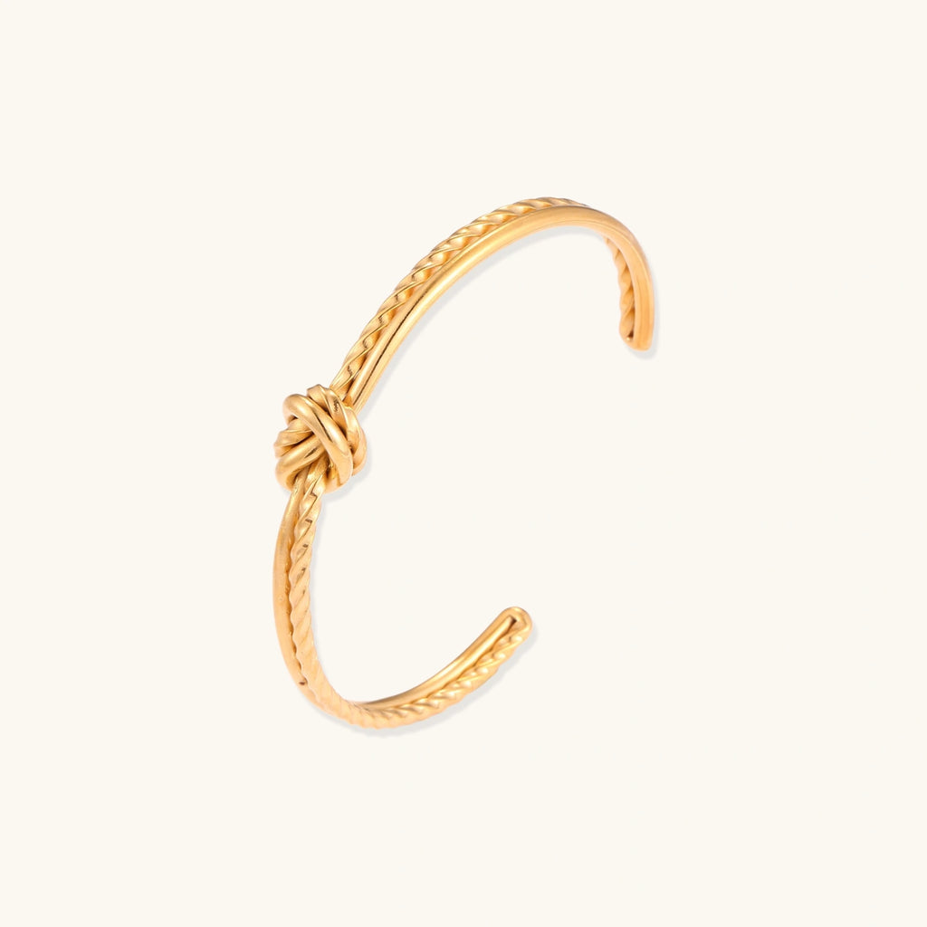 Gold cuff bracelet with twisted rope texture and decorative knot centerpiece on cream background.